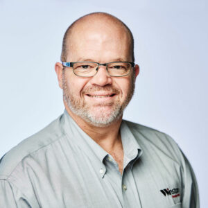 Professional headshot of a smiling man with glasses, wearing a collared shirt, against a light background. This image represents a corporate or business setting.