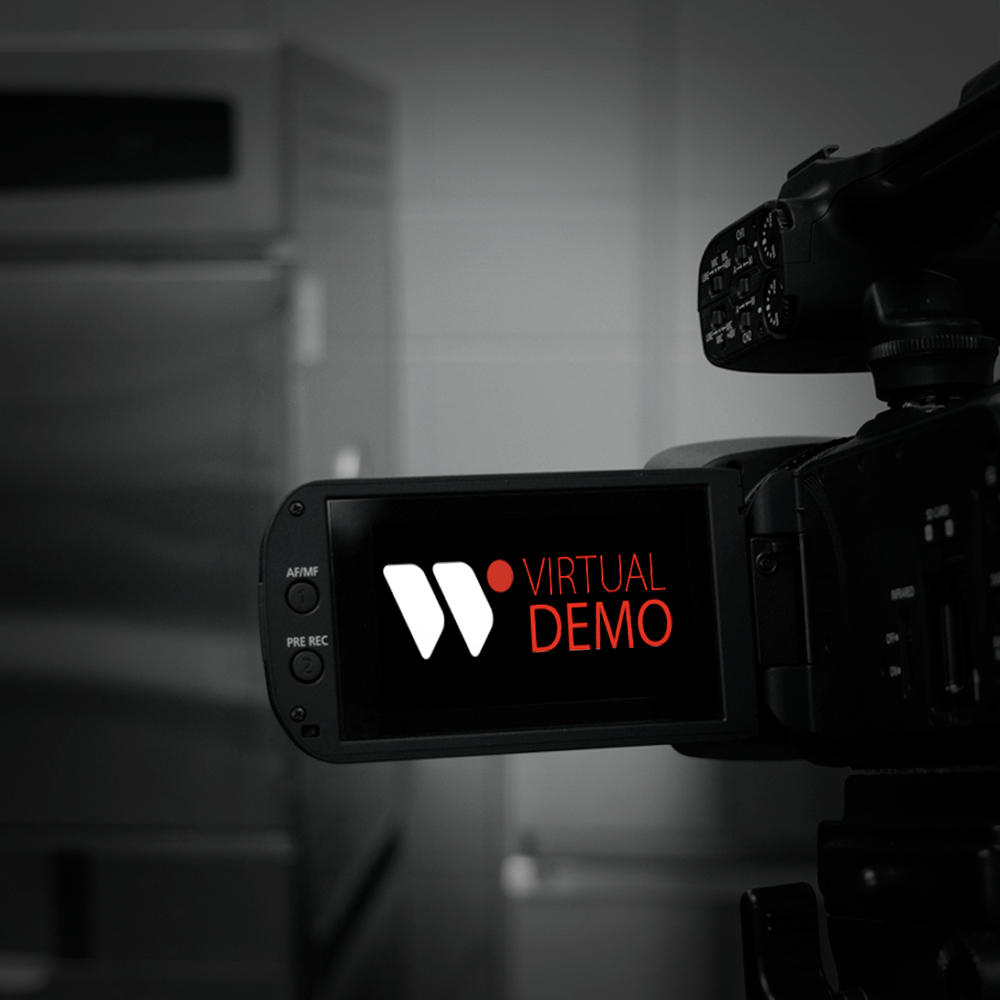 Close-up of a video camera displaying the text "Virtual Demo" on its screen, set against a blurred background of a kitchen or storage area, highlighting video production and demonstration capabilities.