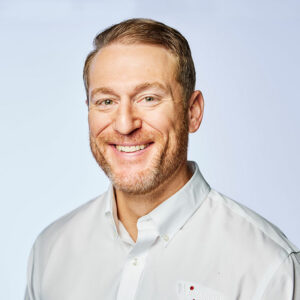 Smiling man in a white shirt against a light background, representing professionalism and approachability in a corporate or medical setting.