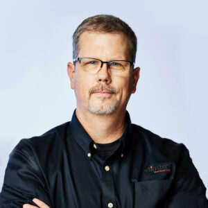 Professional man in glasses wearing a black shirt with arms crossed, presenting a confident and approachable demeanor.