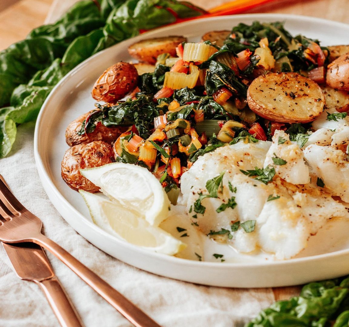 A delicious plate of breakfast featuring poached eggs, sautéed spinach, roasted potatoes, and a colorful vegetable medley, garnished with fresh herbs. The meal is served on a neutral-colored plate with golden utensils, surrounded by fresh spinach leaves.